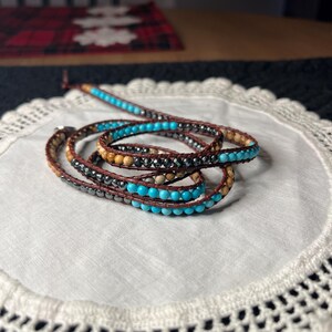 May include: Wrap bracelet featuring turquoise, brown, and black beads. The bracelet is made of multiple strands of beads and leather, creating a layered effect. The beads are a mix of round and cylindrical shapes, adding texture and visual interest to the jewelry.