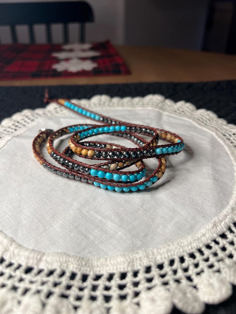 May include: Wrap bracelet featuring turquoise, brown, and black beads. The bracelet is made of multiple strands of beads and leather, creating a layered effect. The beads are a mix of round and cylindrical shapes, adding texture and visual interest to the jewelry.