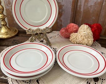 May include: A set of white ceramic plates with a red rim. One plate is displayed on a decorative stand, while others are stacked on a lace doily. The plates have a classic design, suitable for serving meals.