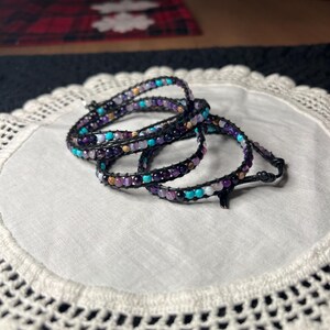 May include: Wrap bracelets with a black leather cord and colorful beads in shades of purple, turquoise, and cream. The bracelets are displayed on a white, crocheted doily.