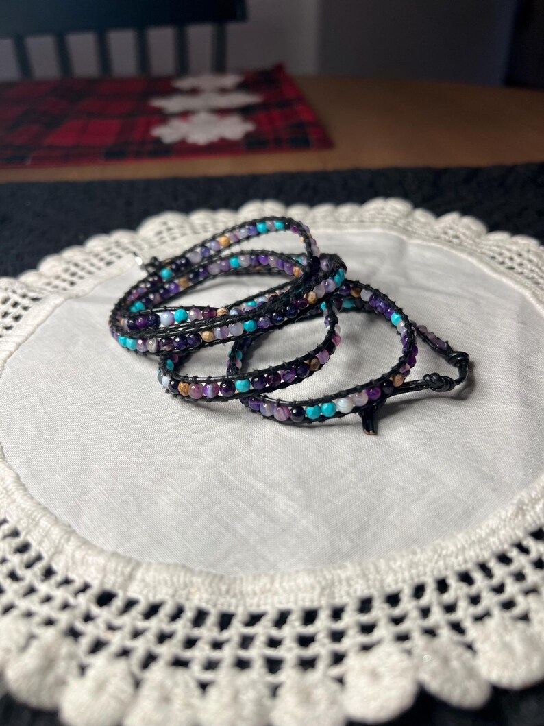 May include: Wrap bracelets with a black leather cord and colorful beads in shades of purple, turquoise, and cream. The bracelets are displayed on a white, crocheted doily.