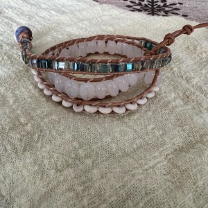 May include: A multi-strand wrap bracelet featuring light brown leather cords and various beads. The bracelet includes clear, white, and iridescent blue-green beads. A silver-toned button clasp secures the bracelet. The bracelet is displayed on a textured, light green surface.