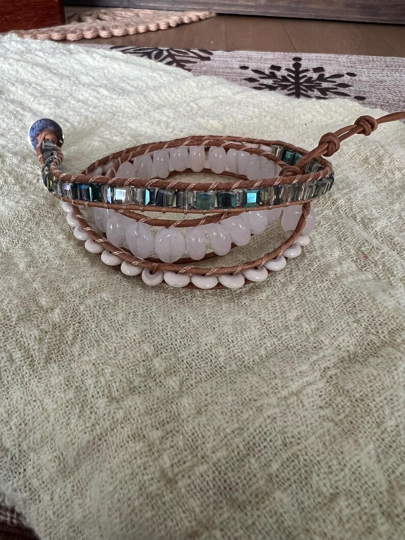 May include: A multi-strand wrap bracelet featuring light brown leather cords and various beads. The bracelet includes clear, white, and iridescent blue-green beads. A silver-toned button clasp secures the bracelet. The bracelet is displayed on a textured, light green surface.
