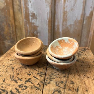 May include: Two stacks of small, terracotta-colored clay bowls. One stack has a natural finish, while the other has a white and orange painted rim. The bowls are on a rustic wooden surface.