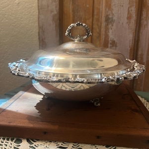 May include: A silver-colored metal serving dish with a matching lid. The dish features a rounded bowl, a decorative scalloped edge with embossed detailing, three small feet, and a central handle on the lid shaped like a floral ring.
