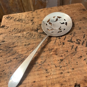 May include: A silver-toned metal serving spoon featuring a wide, circular bowl with a decorative cut-out design of leaves and a central circular element. The handle is smooth, slender, and tapers toward the end, connecting to the round bowl.