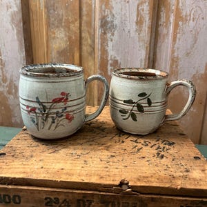 May include: Two off-white ceramic mugs with a speckled texture and brown rim detailing. The left mug features a hand-painted red and blue floral design, while the right mug displays a green leaf motif. Both mugs have horizontal incised lines.