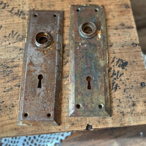 May include: Two antique, rectangular door plates with keyholes and circular knob openings. The metal plates have a weathered, rusty appearance, suggesting age and exposure to the elements. The plates are mounted on a wooden surface.