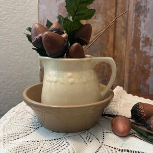 May include: A cream-colored ceramic pitcher with a textured acorn pattern sits inside a tan ceramic bowl. The pitcher is filled with artificial brown acorns and green leaves. Additional brown acorns are placed near the base of the bowl.