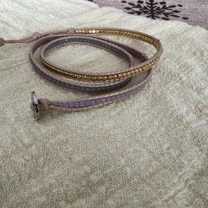 May include: Wrap bracelet with a neutral-coloured cord and a silver clasp. The bracelet features three strands: one with small gold beads, and two with small, square, lavender-coloured beads. The bracelet is wrapped around itself.