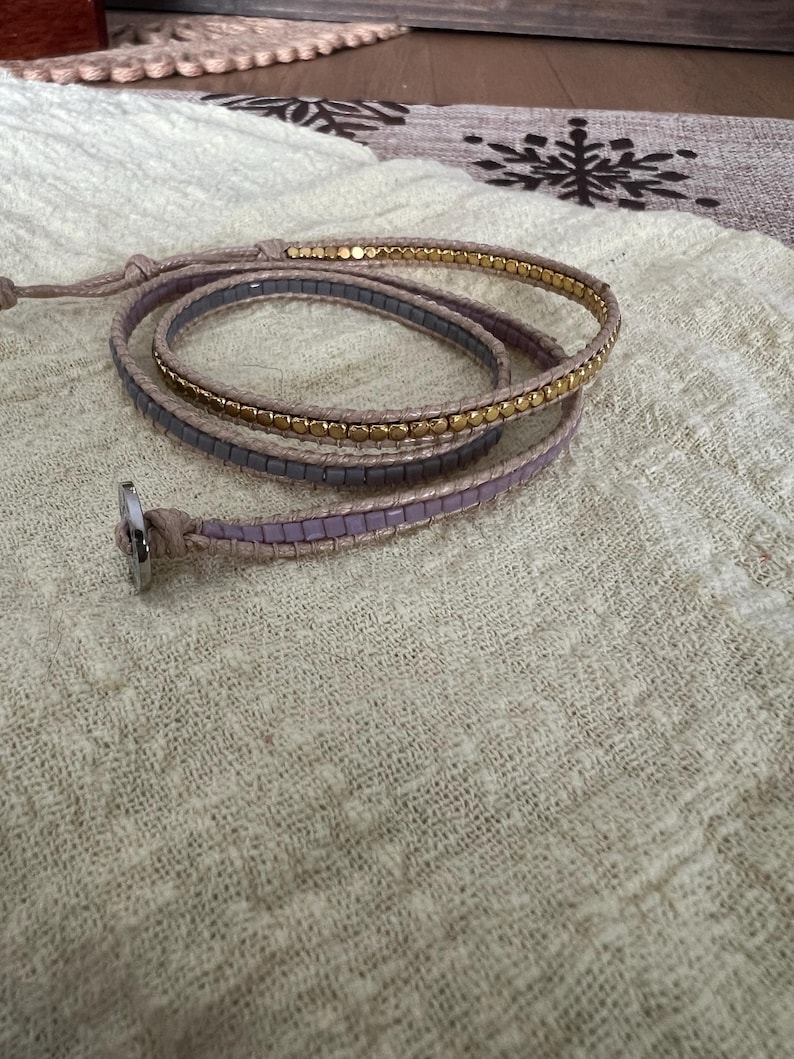 May include: Wrap bracelet with a neutral-coloured cord and a silver clasp. The bracelet features three strands: one with small gold beads, and two with small, square, lavender-coloured beads. The bracelet is wrapped around itself.
