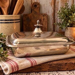May include: A rectangular silver-plated serving dish with a matching lid and two side handles. The lid features a central finial knob and a beaded edge detail. The polished metal surface reflects the surrounding environment.