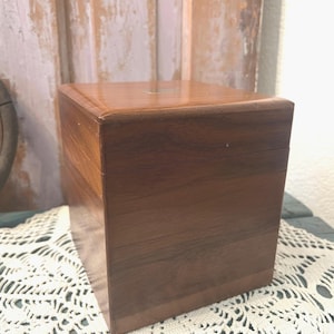 May include: A square wooden box with a smooth, polished finish and visible dark brown wood grain. The box features a flat lid with a small, rectangular metallic plate centered on the top surface. The construction consists of horizontal wood panels.