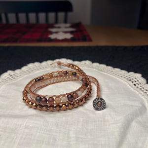 Puede incluir: Pulsera envolvente con m&uacute;ltiples hilos de cuentas en tonos marr&oacute;n, beige y cobre. La pulsera est&aacute; colocada sobre un pa&ntilde;o blanco con bordes de encaje. Un peque&ntilde;o dije redondo con texto est&aacute; adjunto.