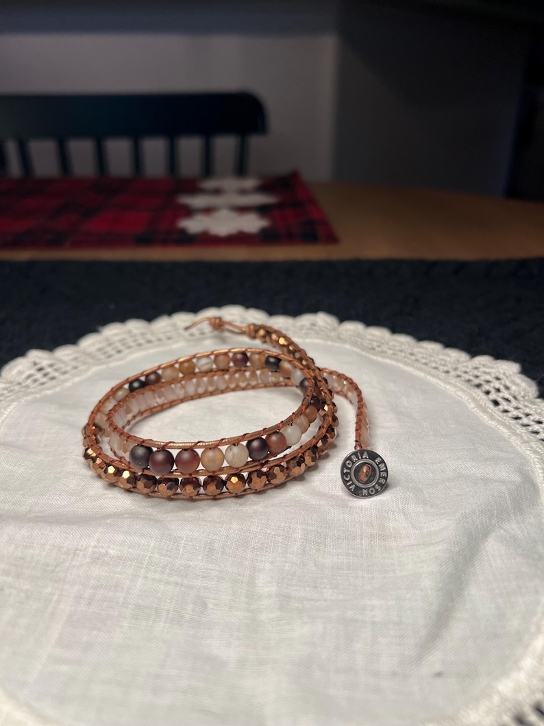 Puede incluir: Pulsera envolvente con m&uacute;ltiples hilos de cuentas en tonos marr&oacute;n, beige y cobre. La pulsera est&aacute; colocada sobre un pa&ntilde;o blanco con bordes de encaje. Un peque&ntilde;o dije redondo con texto est&aacute; adjunto.