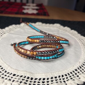 May include: Wrap bracelet featuring turquoise, brown, and silver beads. The bracelet is made with leather and has a wrap-around design. The beads are strung together, creating a layered effect. The bracelet is displayed on a white doily.