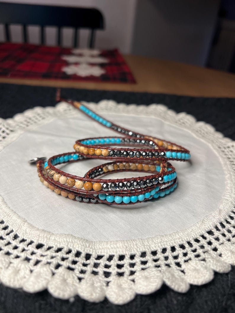 May include: Wrap bracelet featuring turquoise, brown, and silver beads. The bracelet is made with leather and has a wrap-around design. The beads are strung together, creating a layered effect. The bracelet is displayed on a white doily.