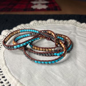 May include: Wrap bracelets with turquoise, brown, and silver beads. The bracelets are made of leather and are wrapped multiple times. The bracelets are on a white, lace-edged surface.