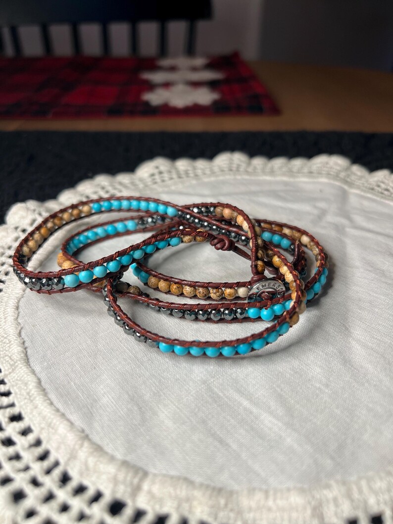 May include: Wrap bracelets with turquoise, brown, and silver beads. The bracelets are made of leather and are wrapped multiple times. The bracelets are on a white, lace-edged surface.