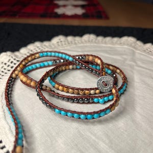 May include: Wrap bracelet with turquoise, brown, and gray beads. The bracelet is made of leather and features a button clasp. The beads are strung on a brown leather cord, creating a layered effect. The bracelet is on a white lace surface.