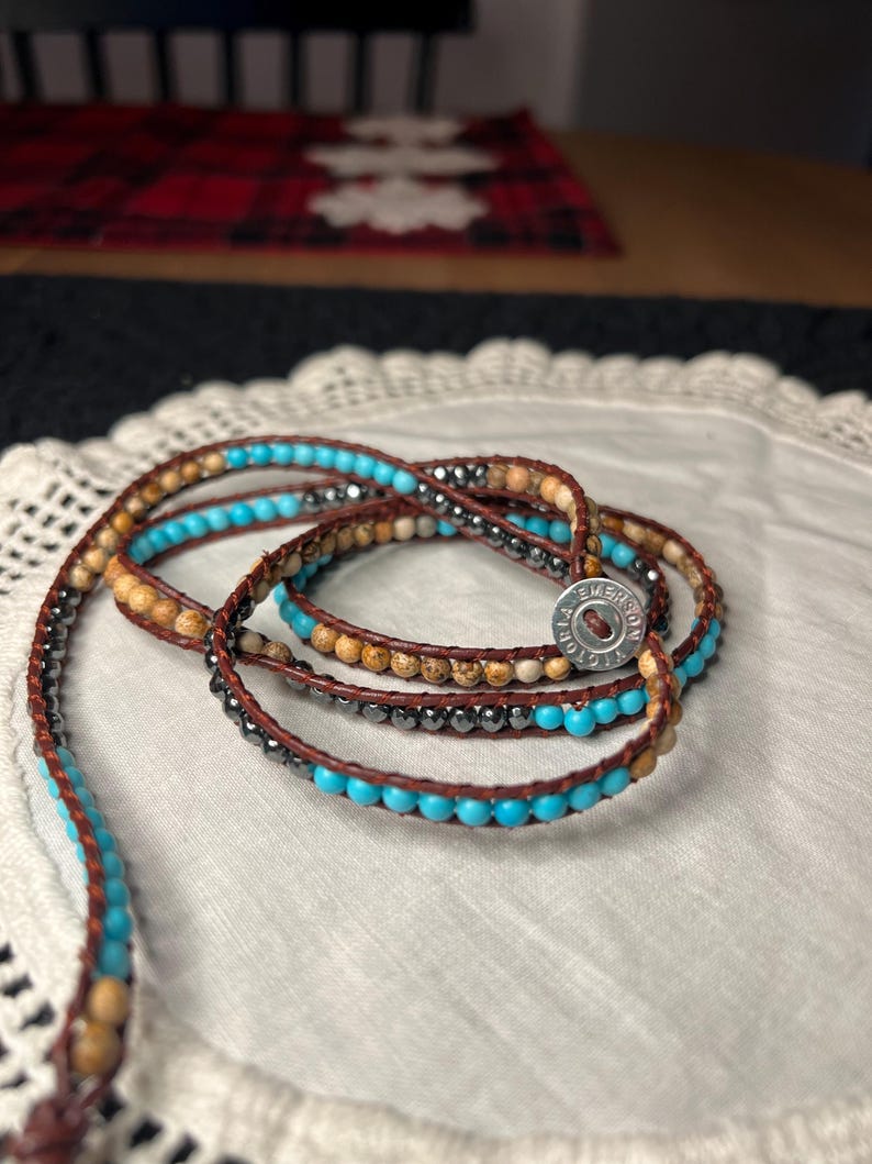 May include: Wrap bracelet with turquoise, brown, and gray beads. The bracelet is made of leather and features a button clasp. The beads are strung on a brown leather cord, creating a layered effect. The bracelet is on a white lace surface.