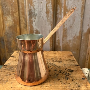 May include: A copper coffee pot with a wide, tapered base and a long, slender handle featuring an engraved scrollwork pattern. The interior of the pot is lined with a silver-colored metal, and the exterior has a polished, reflective copper finish.