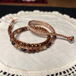 Puede incluir: Una pulsera envolvente con alambre de cobre y varias cuentas en tonos marr&oacute;n, beige y transparente. La pulsera est&aacute; enrollada sobre un pa&ntilde;o blanco con bordes de encaje. El cierre es un peque&ntilde;o bot&oacute;n plateado.