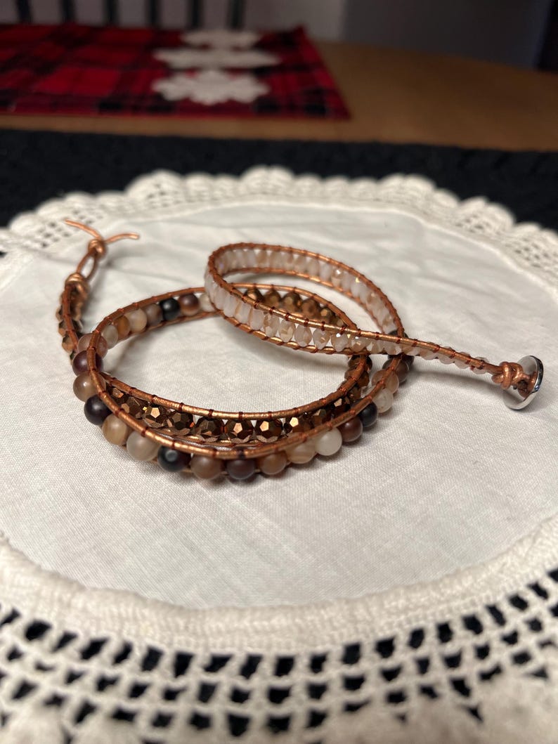 Puede incluir: Una pulsera envolvente con alambre de cobre y varias cuentas en tonos marr&oacute;n, beige y transparente. La pulsera est&aacute; enrollada sobre un pa&ntilde;o blanco con bordes de encaje. El cierre es un peque&ntilde;o bot&oacute;n plateado.