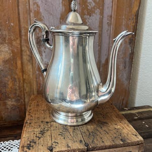 May include: A silver-colored coffee pot with a curved spout and handle. The lid is topped with a decorative finial. The pot sits on a wooden crate with the text "REMINGTON ARMS COMPANY, INC." and "BRIDGEPORT WORKS" visible.