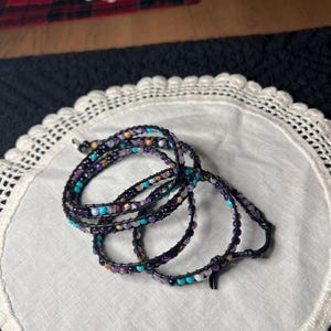 May include: Wrap bracelet with a black cord and colorful beads in shades of purple, turquoise, and gold. The bracelet is wrapped multiple times and is resting on a white, lace-edged surface.