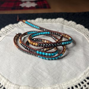 May include: Wrap bracelet featuring turquoise, brown, and silver beads. The bracelet is made with a brown leather cord and wraps multiple times around the wrist. The beads are arranged in a repeating pattern.