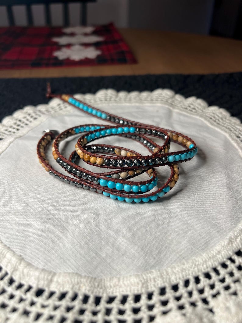 May include: Wrap bracelet featuring turquoise, brown, and silver beads. The bracelet is made with a brown leather cord and wraps multiple times around the wrist. The beads are arranged in a repeating pattern.