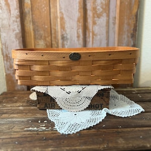 May include: A rectangular, light brown woven basket with a metal emblem. The basket is set on a wooden crate with the text "AIR SHOT" visible. A white, lacy doily is draped over the crate and basket, creating a vintage aesthetic.