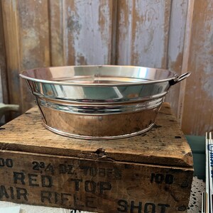 May include: A shiny, silver-colored metal basin with a rolled rim and small handles. The basin is round and sits on a wooden box with faded black text. The background is a weathered wooden wall.