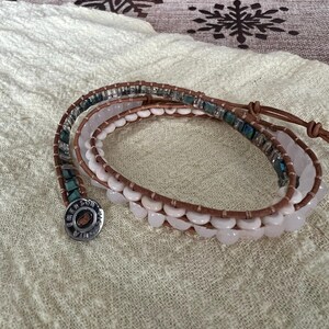 May include: Wrap bracelet featuring a combination of light brown leather, white and pink beads, and turquoise accents. The bracelet has a silver-toned button clasp. The design is suitable for everyday wear.