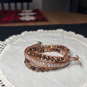 Puede incluir: Pulsera envolvente con m&uacute;ltiples hilos de cuentas en tonos marr&oacute;n, beige y transparente, ensartadas en alambre de cobre. La pulsera est&aacute; enrollada sobre s&iacute; misma y asegurada con un cierre de alambre de cobre. La pulsera est&aacute; sobre una superficie blanca y de encaje.