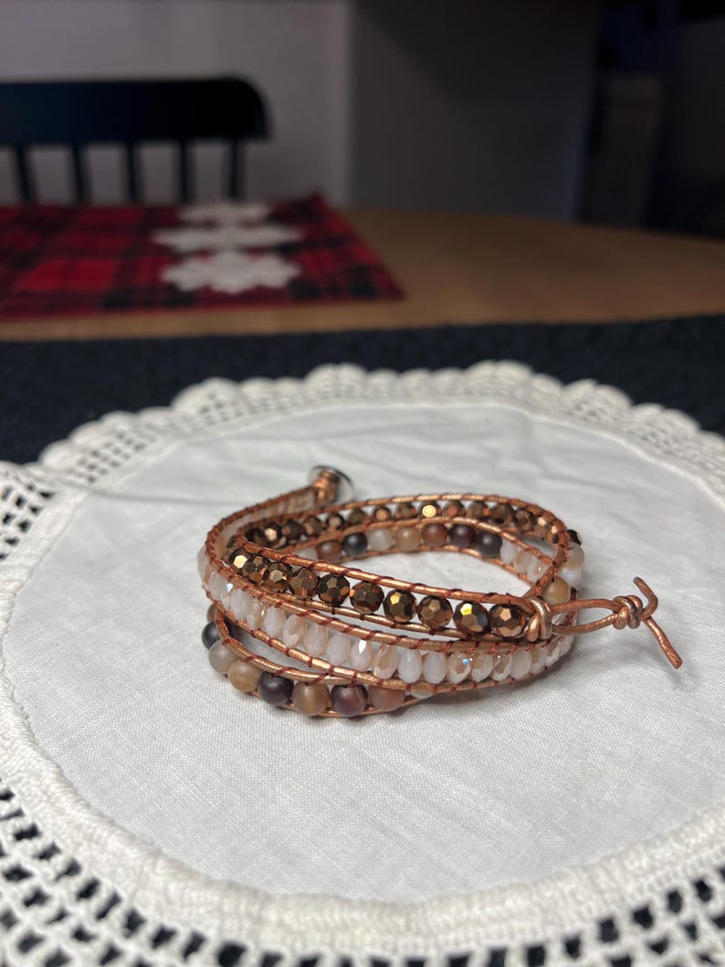 Puede incluir: Pulsera envolvente con m&uacute;ltiples hilos de cuentas en tonos marr&oacute;n, beige y transparente, ensartadas en alambre de cobre. La pulsera est&aacute; enrollada sobre s&iacute; misma y asegurada con un cierre de alambre de cobre. La pulsera est&aacute; sobre una superficie blanca y de encaje.