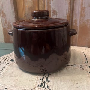 May include: A dark brown ceramic pot with a matching lid and two small side handles. The pot features a smooth, glossy finish and a cylindrical body shape. The lid has a small, centered knob handle on top, and the overall design is simple.