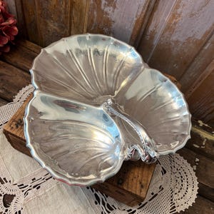 May include: A silver-toned metal serving dish shaped like a three-sectioned shell. The dish features a central handle with ornate detailing and a scalloped edge design. The surface is polished and reflective, showing a smooth, curved interior.