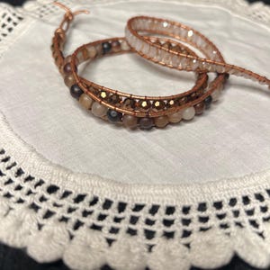 Puede incluir: Una pulsera envolvente hecha a mano con un dise&ntilde;o de alambre de cobre y varias cuentas de tonos neutros. La pulsera est&aacute; enrollada sobre s&iacute; misma, mostrando el intrincado trabajo de cuentas y los tonos c&aacute;lidos. La pulsera est&aacute; sobre un tapete de encaje blanco.