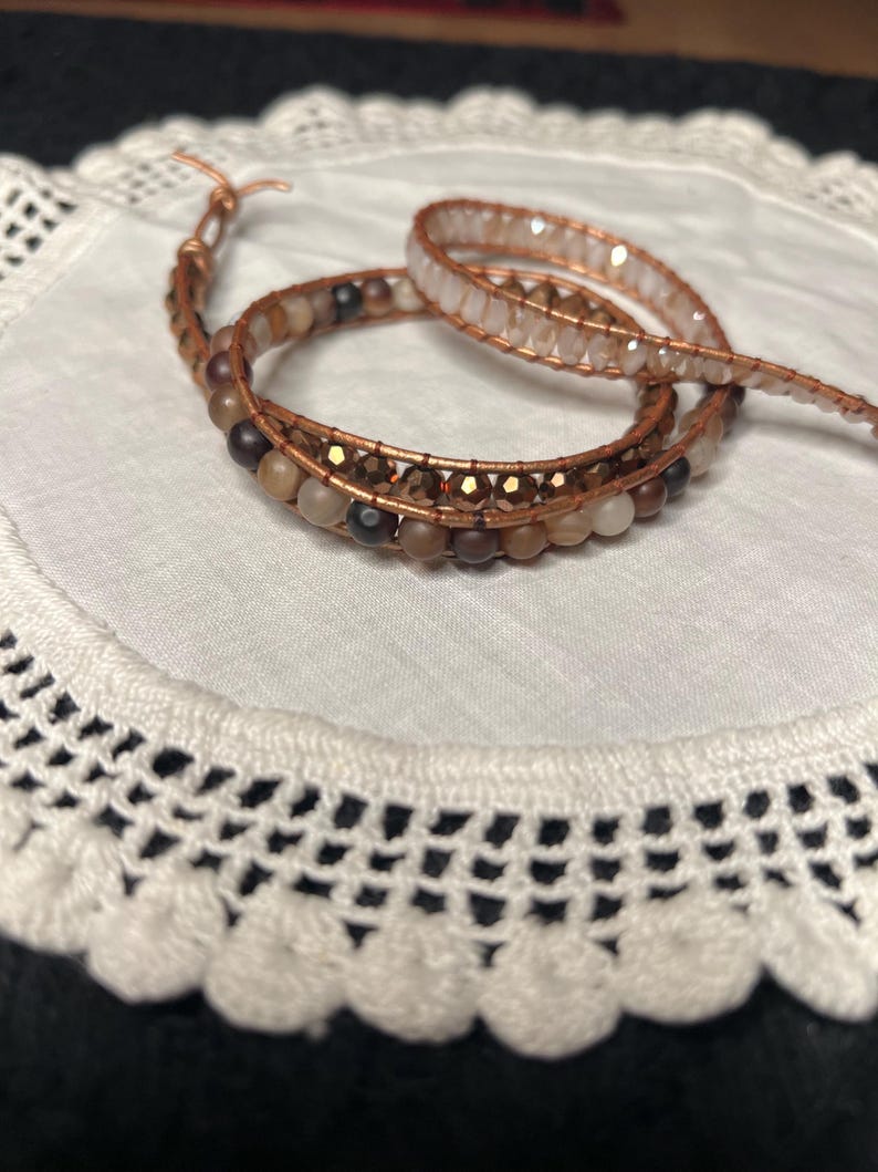 Puede incluir: Una pulsera envolvente hecha a mano con un dise&ntilde;o de alambre de cobre y varias cuentas de tonos neutros. La pulsera est&aacute; enrollada sobre s&iacute; misma, mostrando el intrincado trabajo de cuentas y los tonos c&aacute;lidos. La pulsera est&aacute; sobre un tapete de encaje blanco.