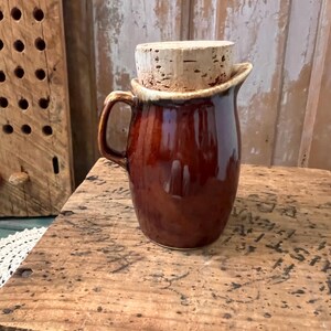 May include: A brown ceramic pitcher with a cork stopper. The pitcher has a handle and a wide rim with a lighter brown glaze. The cork stopper is cylindrical and fits snugly in the pitcher's opening. The pitcher is sitting on a wooden surface.