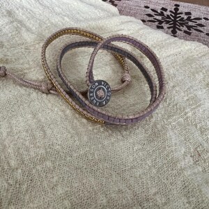 May include: A wrap bracelet with a light brown leather band and a gold chain accent. The bracelet features a button clasp with the text "SON VICTORIA EMERSON". The bracelet is lying on a cream-coloured fabric.