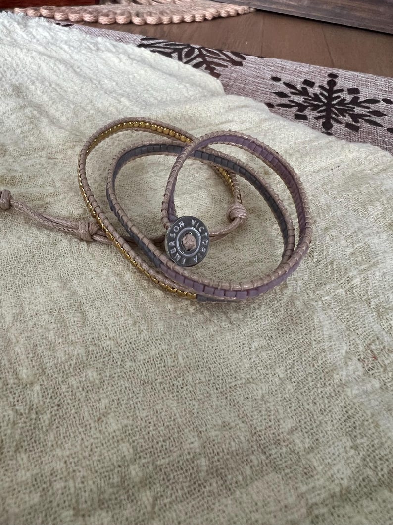 May include: A wrap bracelet with a light brown leather band and a gold chain accent. The bracelet features a button clasp with the text "SON VICTORIA EMERSON". The bracelet is lying on a cream-coloured fabric.