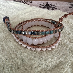 May include: Wrap bracelet featuring light brown leather strands, adorned with rows of white, light pink, and teal-colored beads. The bracelet has a button closure and is designed to wrap multiple times around the wrist. The beads are of varying shapes and sizes.