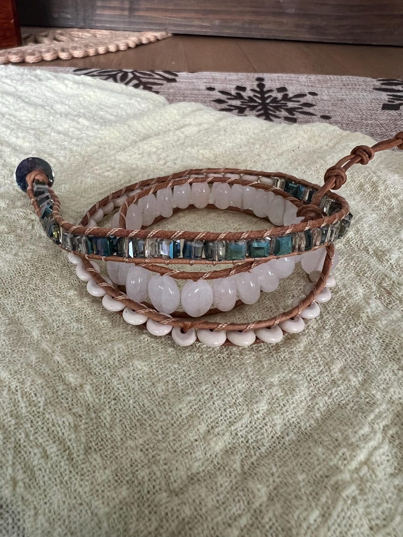 May include: Wrap bracelet featuring light brown leather strands, adorned with rows of white, light pink, and teal-colored beads. The bracelet has a button closure and is designed to wrap multiple times around the wrist. The beads are of varying shapes and sizes.