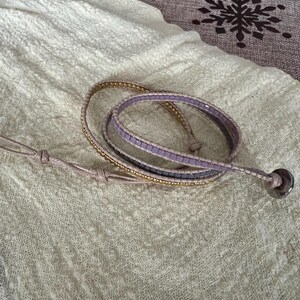 May include: A wrap bracelet with a light purple and gold beaded design. The bracelet is made of multiple strands and features a silver-coloured button clasp. The bracelet is lying on a cream-coloured fabric.