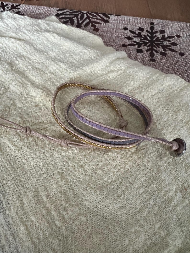 May include: A wrap bracelet with a light purple and gold beaded design. The bracelet is made of multiple strands and features a silver-coloured button clasp. The bracelet is lying on a cream-coloured fabric.