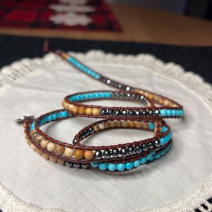 May include: Wrap bracelet featuring brown leather and various beads. The bracelet includes turquoise, brown, and black beads. The design is a multi-strand wrap style, suitable for a bohemian or casual aesthetic.