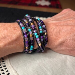 May include: Wrap bracelet featuring a multi-strand design with purple, turquoise, and tan beads. The bracelet is strung on black cord and wraps multiple times around the wrist. The beads are various sizes and shapes.